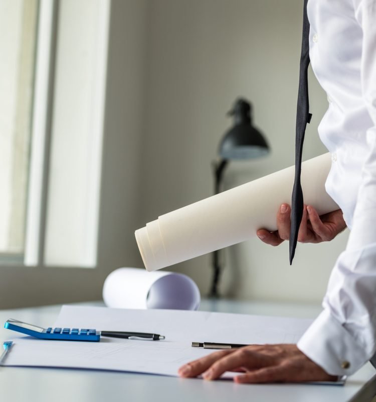 Architect or engineer leaning on his office desk holding rolled blueprints in his hands and leaning over another project on the table.